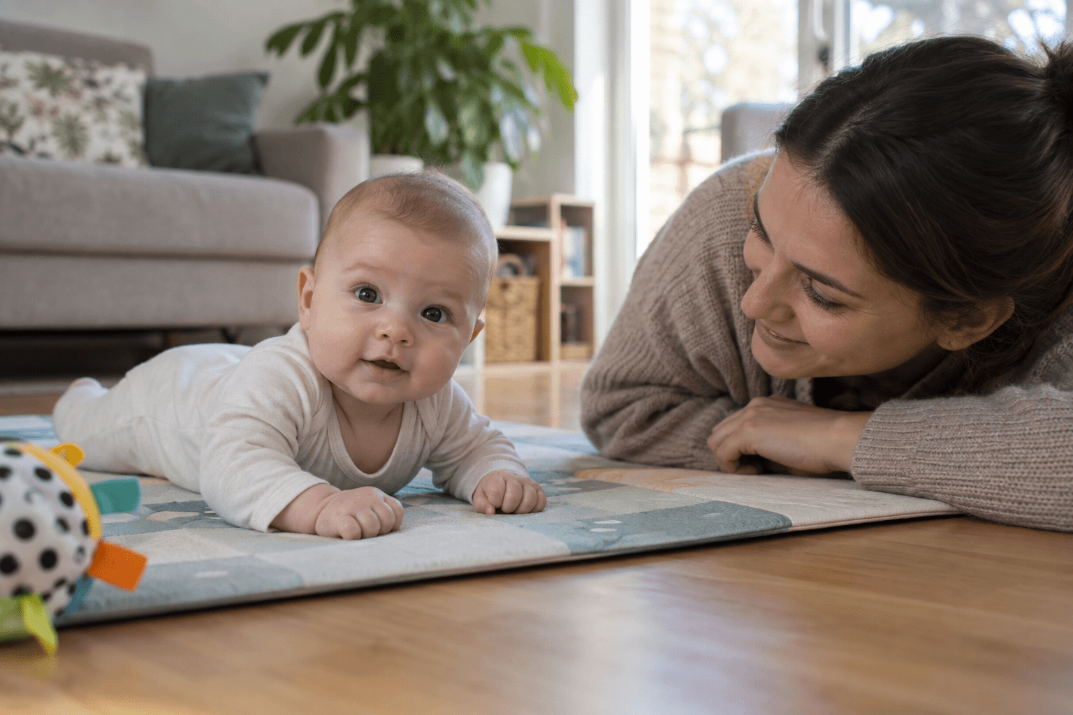 Bebeluș treaz sprijinit pe antebrațe pe o salteluță de joacă, cu mama culcată alături și atentă la el.