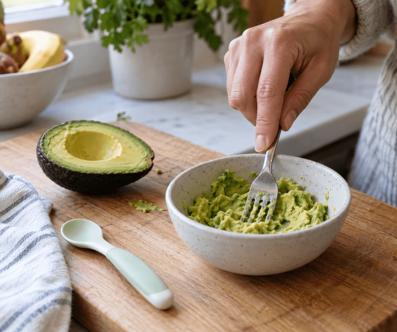 Avocado pasat cu furculița într-un bol mic, lângă o linguriță și o jumătate de fruct pe un tocător.