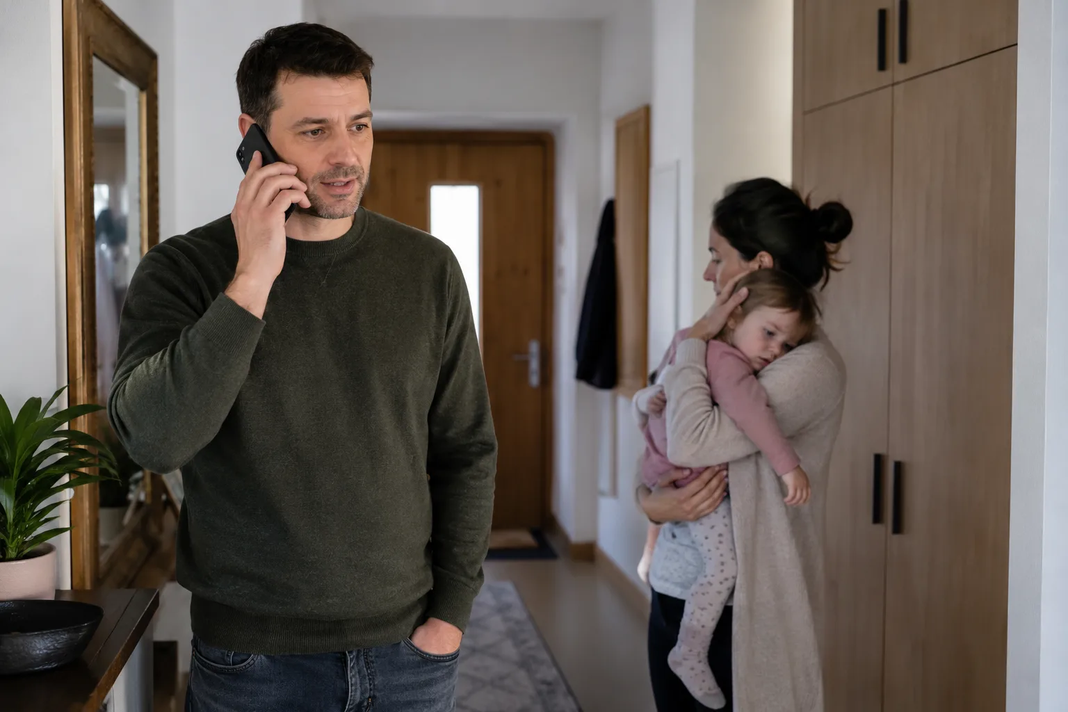 Părinte care sună calm pentru ajutor în locuință, cu un copil mic ținut de un alt adult, fără text sau logo