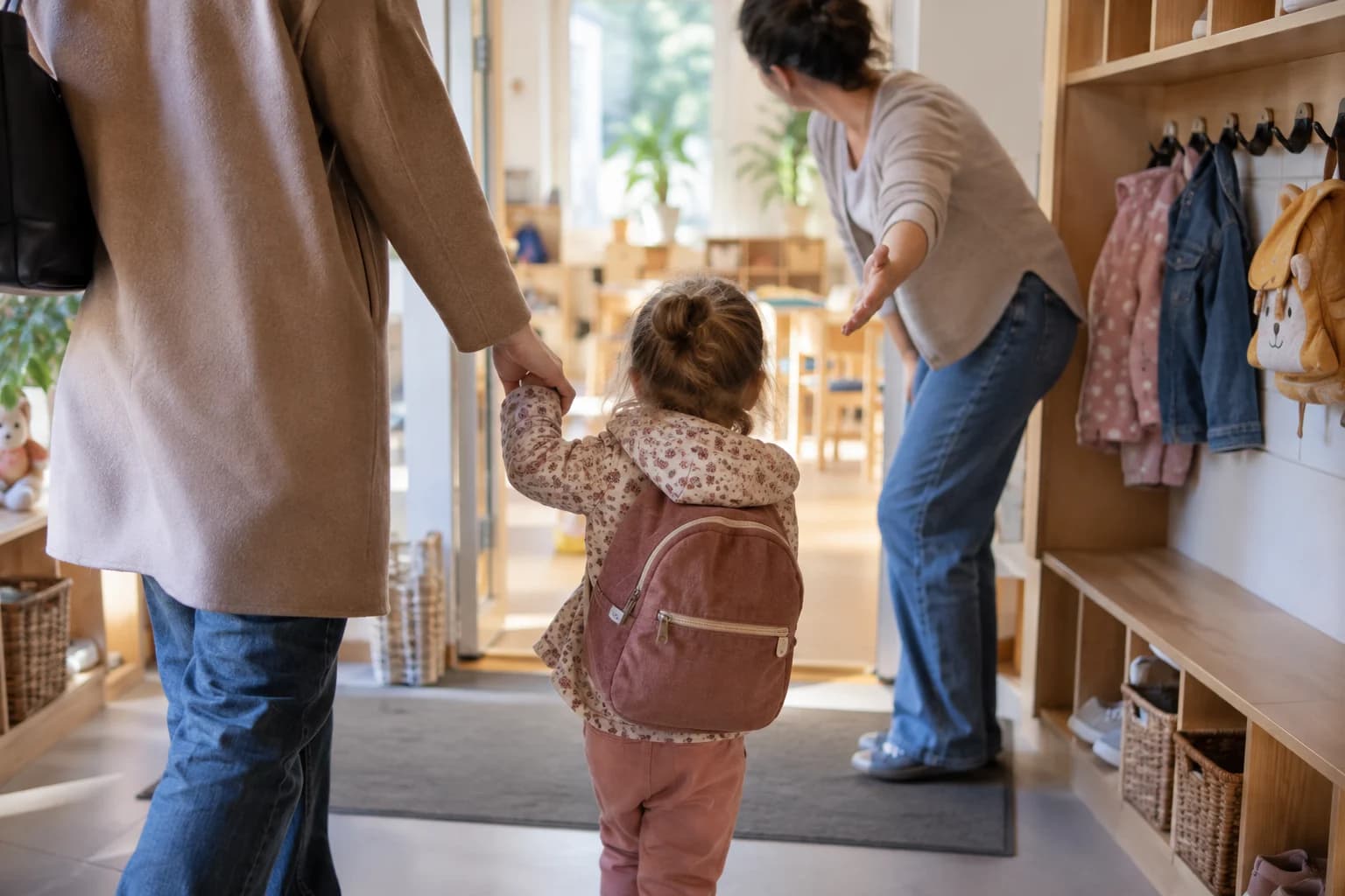 Copil mic la intrarea într-o creșă, ținând mâna părintelui și salutând o educatoare, fără text sau logo